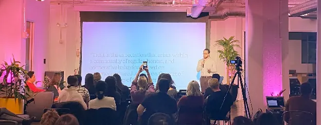 A speaker on stage at a Magenta Talks event in Brooklyn, with an audience seated in front of a magenta-lit backdrop.